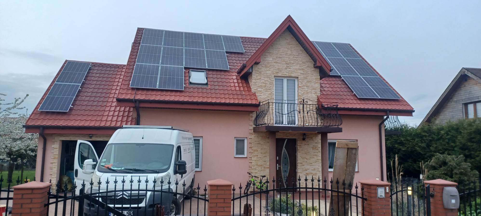 Dom jednorodzinny z czerwonym dachem i panelami słonecznymi, widoczny zza czarnego, kutego ogrodzenia. Obok domu zaparkowany biały bus z otwartymi drzwiami.