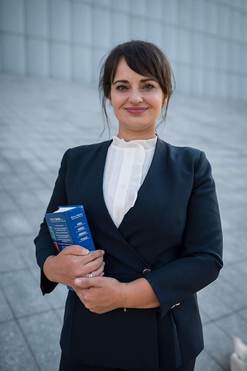 Uśmiechnięta kobieta w ciemnym garniturze i białej bluzce z falbanami, trzymająca w rękach niebieską książkę z napisem 'Edycja Sądowa' i skrótami KSH, KRSU, MSiGU, PrUpad, na tle jasnej...