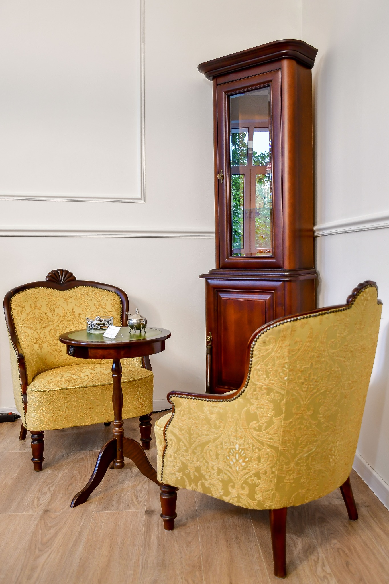 Antyczne fotele z wzorzystą tapicerką, stolik z dekoracjami i witryna w rogu pokoju. Klasyczny wystrój wnętrza z drewnianą podłogą.