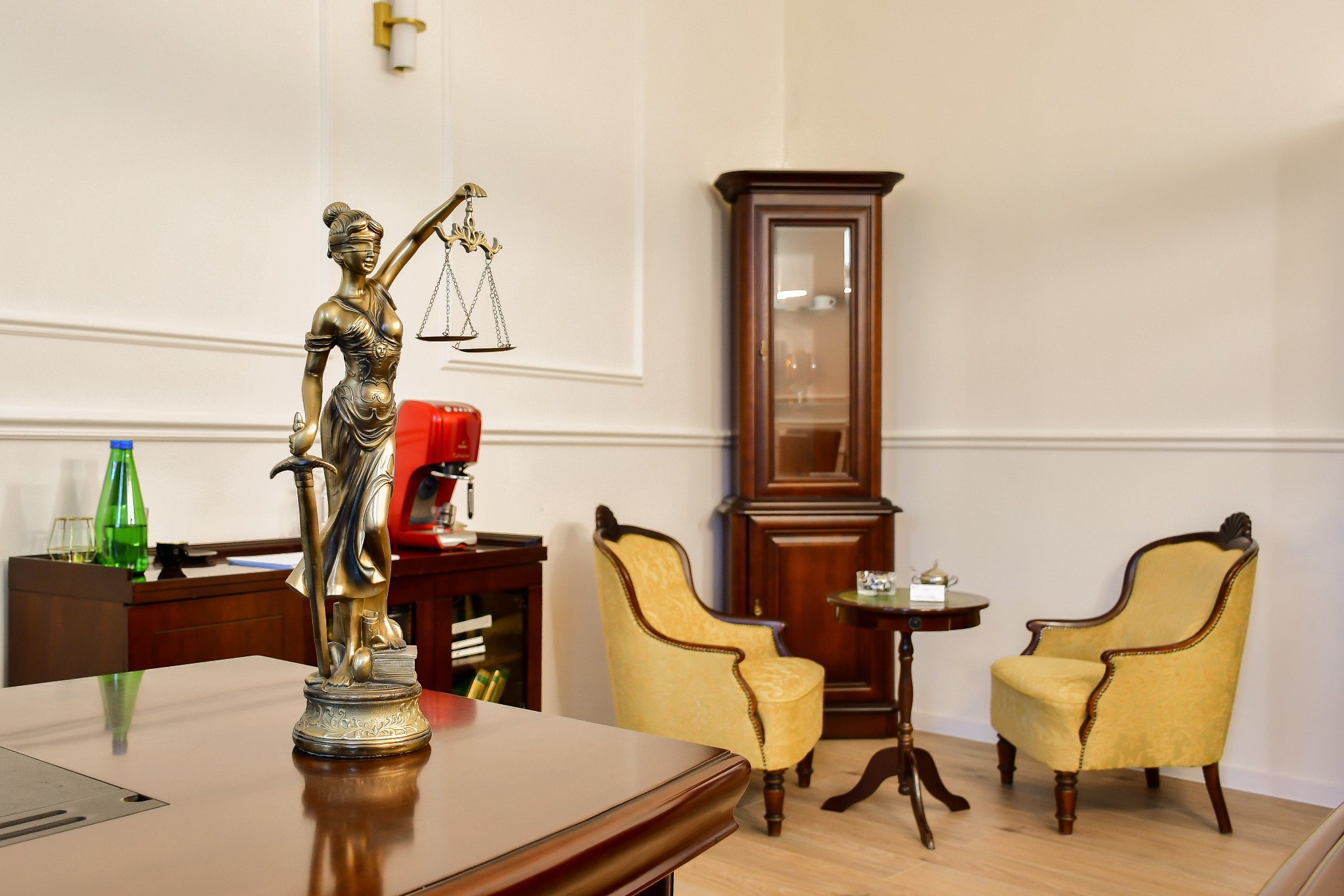 Statuetka Temidy na biurku w kancelarii, w tle krzesła, kredens z ekspozycją i czerwony ekspres do kawy. Eleganckie wnętrze, ciepłe światło.
