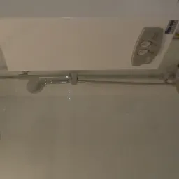 Biały podgrzewacz wody nad kabiną prysznicową z chromowaną armaturą i deszczownicą, widoczne przyłącza i regulatory temperatury.