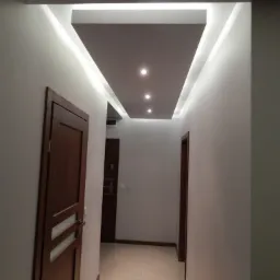 Korytarz z podwieszonym sufitem z oświetleniem LED, widoczne trzy punktowe lampy, brązowe drzwi z przeszkleniami i płytki podłogowe w odcieniach beżu.