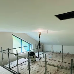 Sufit poddasza w trakcie szlifowania gładzi, widoczne rusztowanie, szlifierka z odkurzaczem, okna zabezpieczone folią i lampa oświetleniowa.