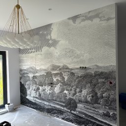 USŁUGI REMONTOWO-BUDOWLANE Łukasz Wojcieszek - Tapetowanie ściany z motywem pejzażu w stylu retro, widoczna nowoczesna lampa sufitowa i narzędzia do tapetowania na podłodze.