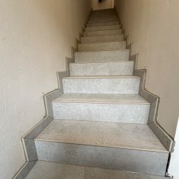 USŁUGI REMONTOWO-BUDOWLANE Łukasz Wojcieszek - Wykończone schody wewnętrzne z szarych płytek ceramicznych, z widocznymi listwami przypodłogowymi i wieszakiem z ubraniem na górze.