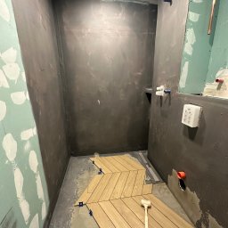 USŁUGI REMONTOWO-BUDOWLANE Łukasz Wojcieszek - Układanie drewnopodobnych płytek w jodełkę w surowej łazience z przygotowanymi ścianami, widoczny gumowy młotek i niebieskie kliny dystansowe.