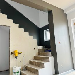 USŁUGI REMONTOWO-BUDOWLANE Łukasz Wojcieszek - Schody w trakcie remontu: ściany częściowo pomalowane na szaro, mikser do gładzi i wiadro z gładzią. Widoczne surowe schody i przygotowane ściany.