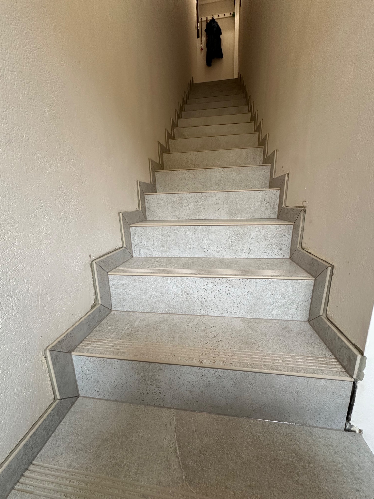 Wykończone schody wewnętrzne z szarych płytek ceramicznych, z widocznymi listwami przypodłogowymi i wieszakiem z ubraniem na górze.