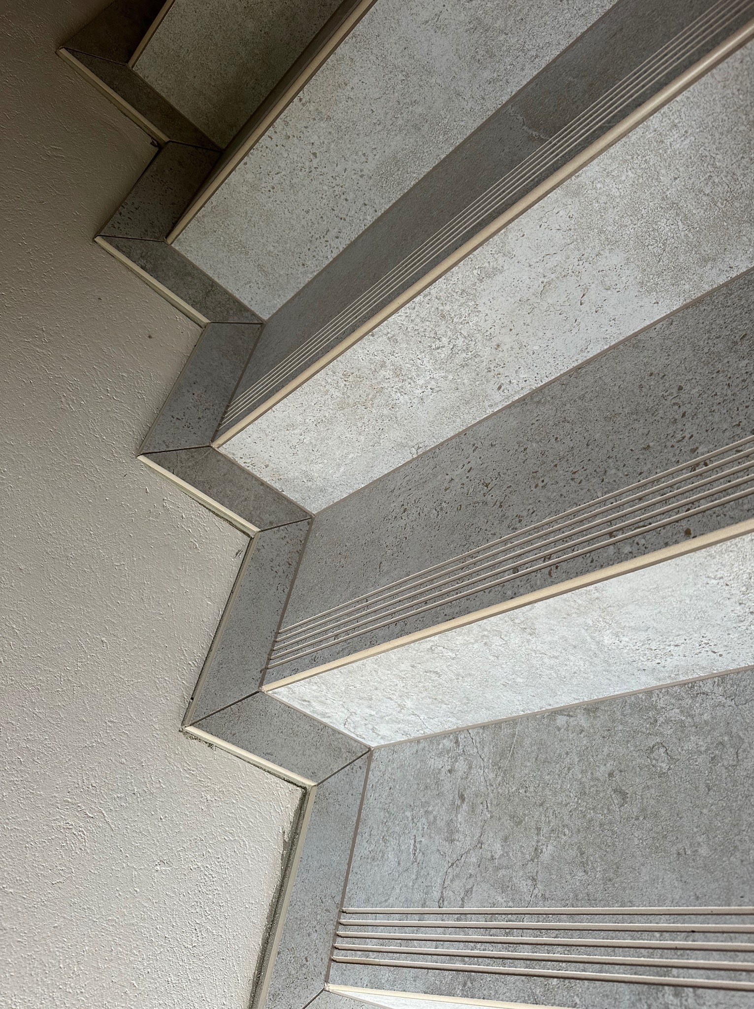 Wykończone schody z szarych płytek imitujących kamień, z metalowymi listwami antypoślizgowymi, widoczne z góry, przylegające do ściany o wyraźnej fakturze.