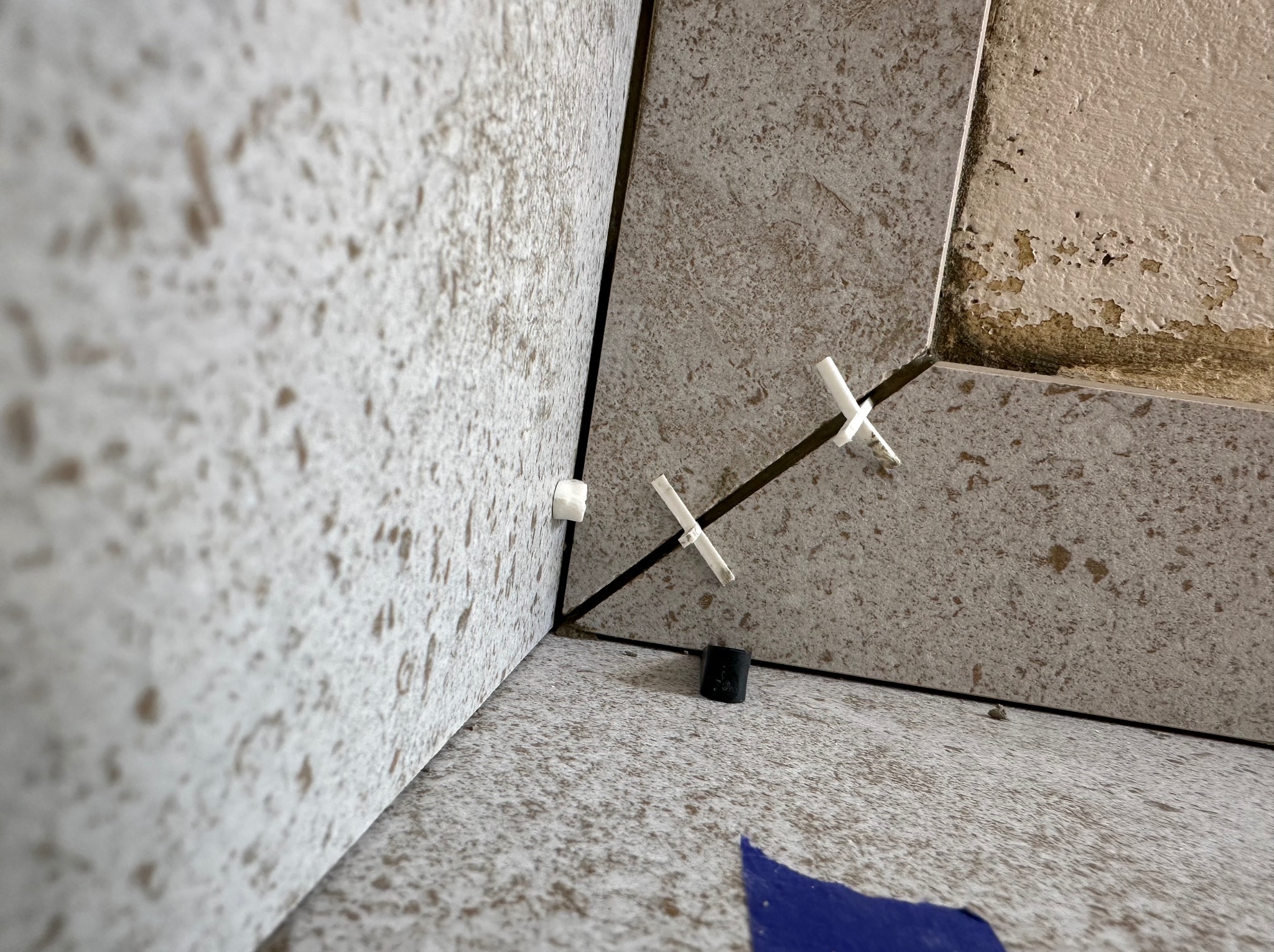 Fragment narożnika z ułożonymi płytkami imitującymi kamień, z krzyżykami dystansowymi i fragmentem niebieskiej taśmy malarskiej na podłodze, widoczne ślady po starej farbie na ścianie.
