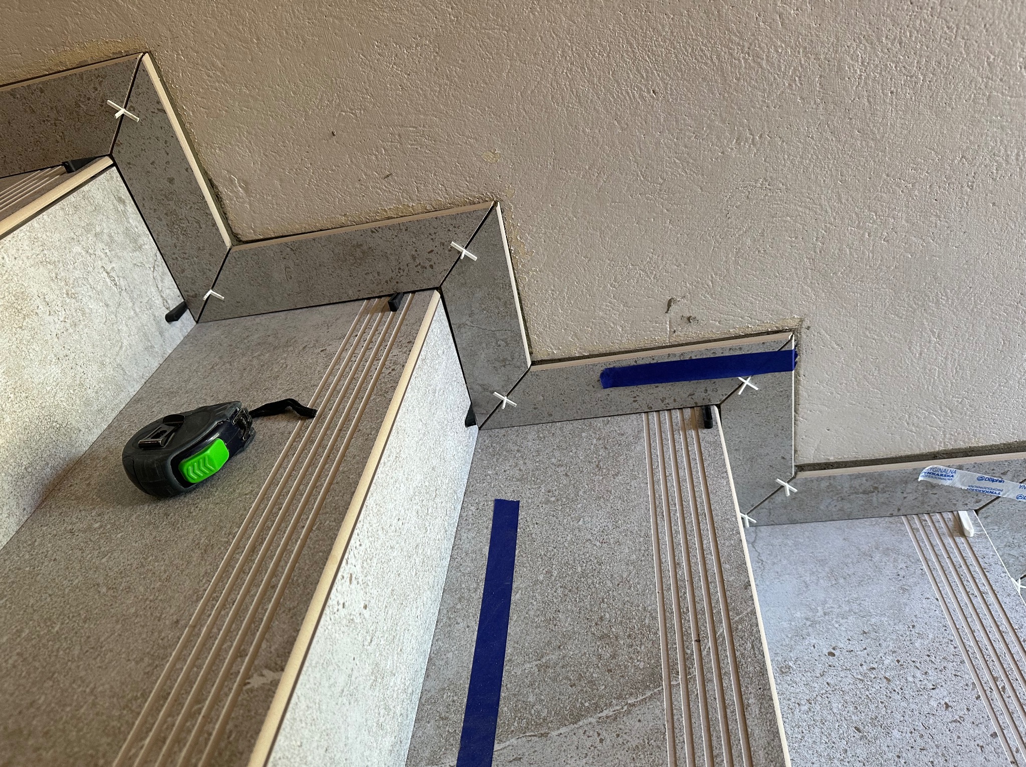 Układanie płytek na schodach z użyciem krzyżyków dystansowych i taśmy maskującej, widoczna miara zwinięta na jednym ze stopni.