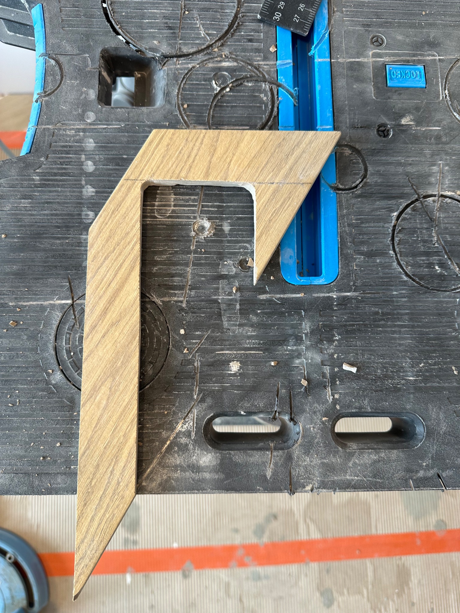 Przycięty element w kształcie litery L z płytek drewnopodobnych, leżący na brudnym stole roboczym z niebieskimi elementami i śladami po cięciu.