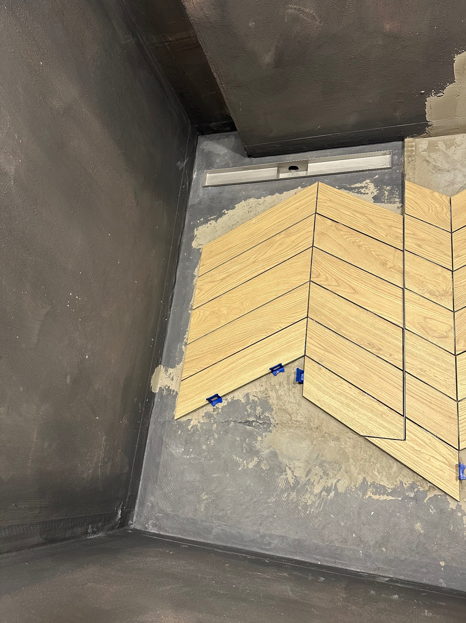 Układanie płytek drewnopodobnych w jodełkę w łazience; widoczny odpływ liniowy i niezagruntowane ściany.