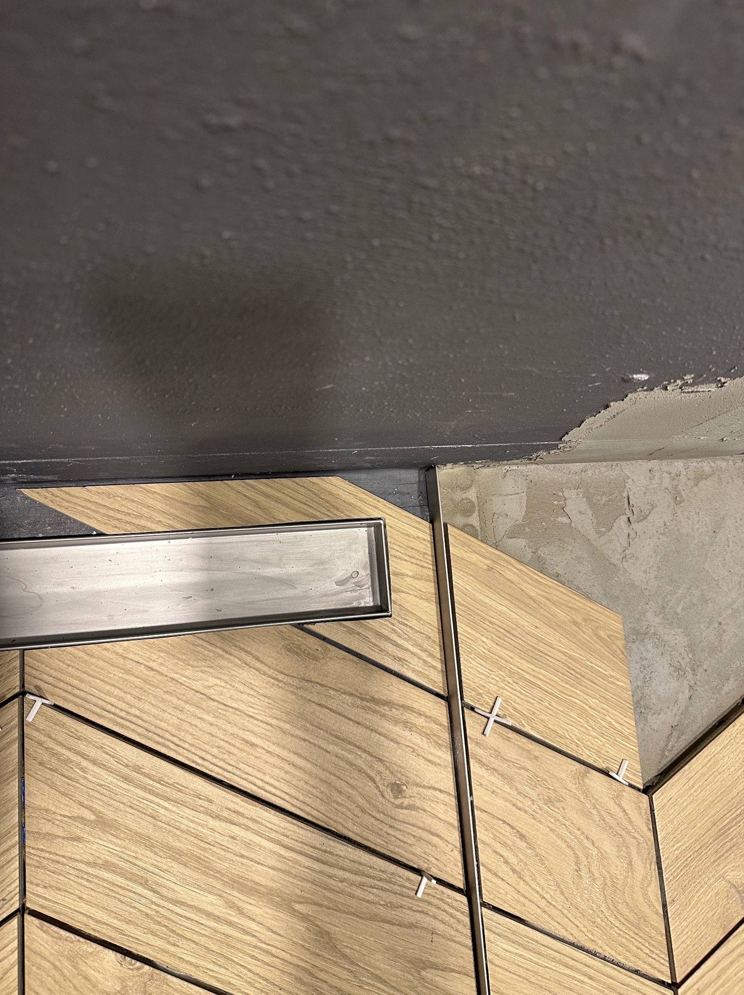 Ułożona w jodełkę jasna okładzina z imitacją drewna w łazience, widoczny odpływ liniowy i krzyżyki dystansowe, surowy sufit w tle.