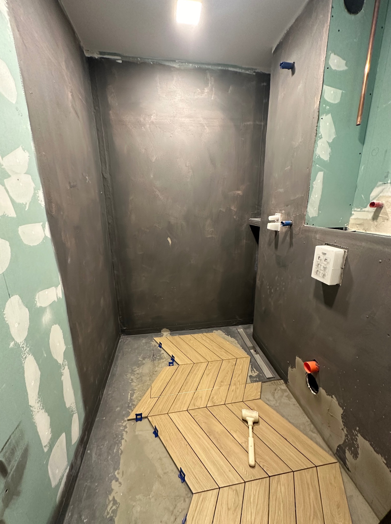 Układanie drewnopodobnych płytek w jodełkę w surowej łazience z przygotowanymi ścianami, widoczny gumowy młotek i niebieskie kliny dystansowe.