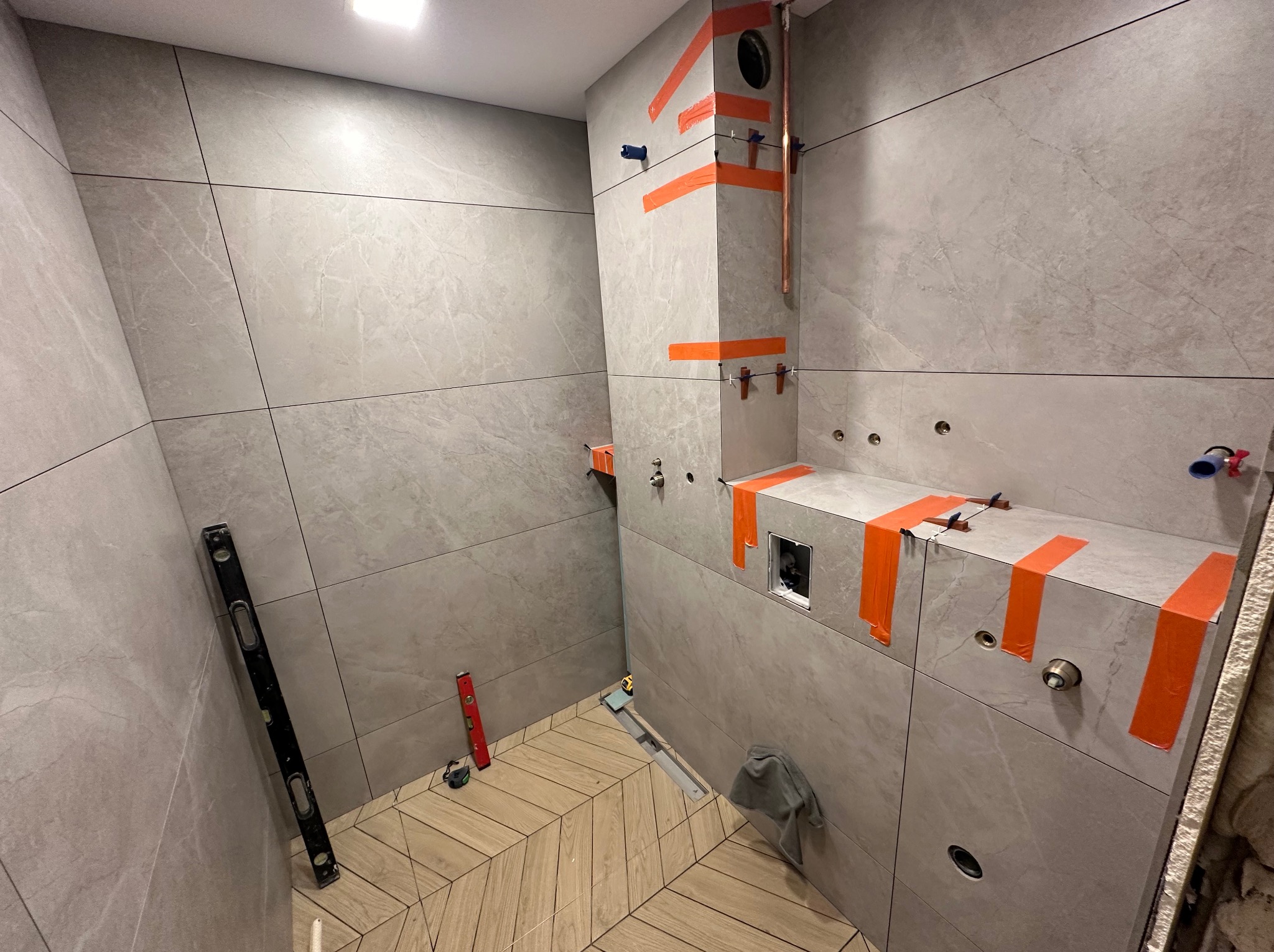 Łazienka w trakcie wykańczania: szare, wielkoformatowe płytki na ścianach, podkreślone czarnymi fugami, fragment podłogi wyłożony parkietem w jodełkę, widoczne narzędzia glazurnicze, rury...