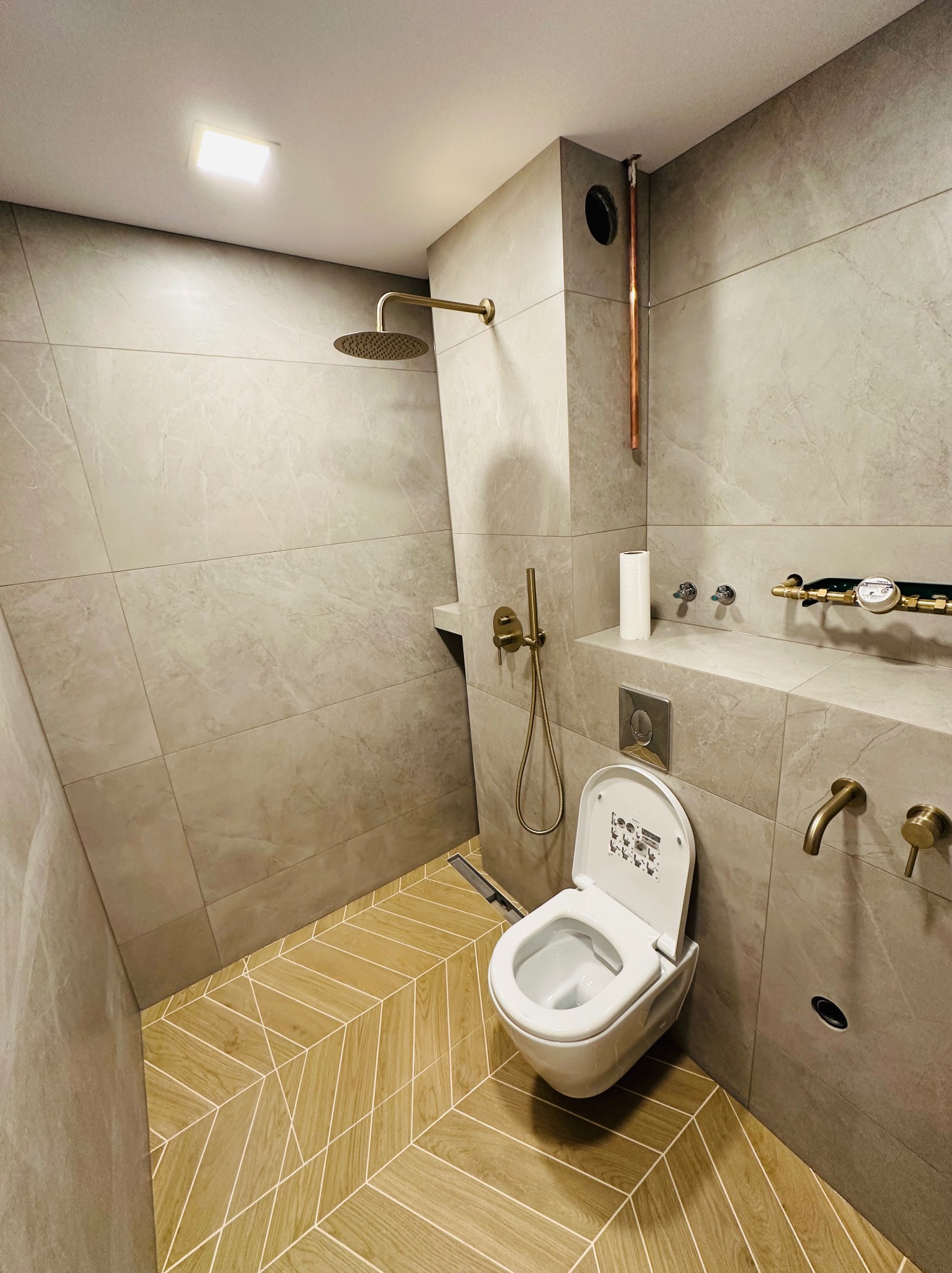 Nowoczesna łazienka z beżowymi płytkami imitującymi marmur, drewnopodobną podłogą ułożoną w jodełkę, złotą armaturą i wiszącą toaletą z otwartą klapą.