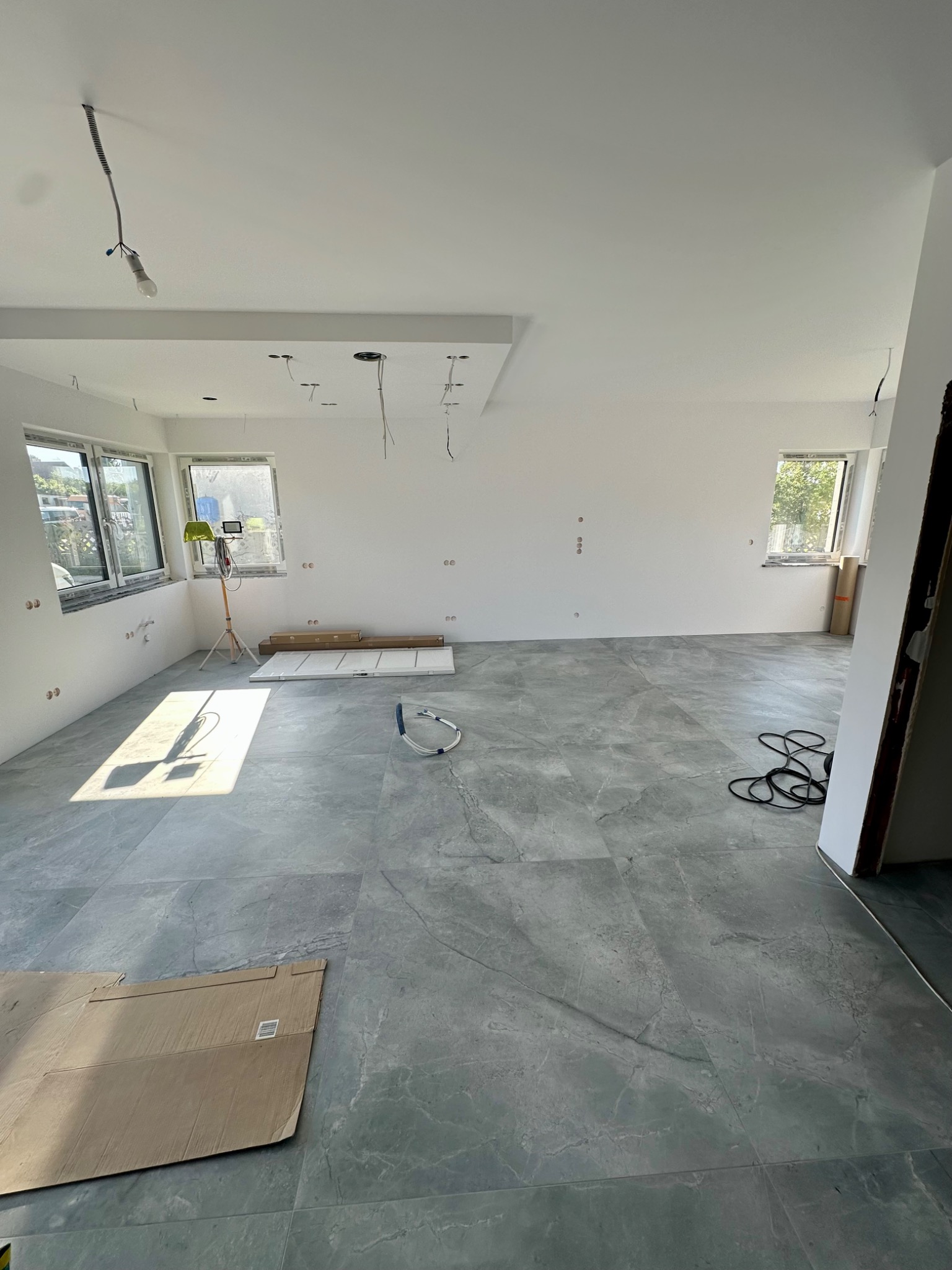 Jasne, przestronne wnętrze z szarą podłogą wyłożoną płytkami imitującymi kamień. Widoczne przygotowania do dalszych prac wykończeniowych, lampa na statywie i rozłożone kable.
