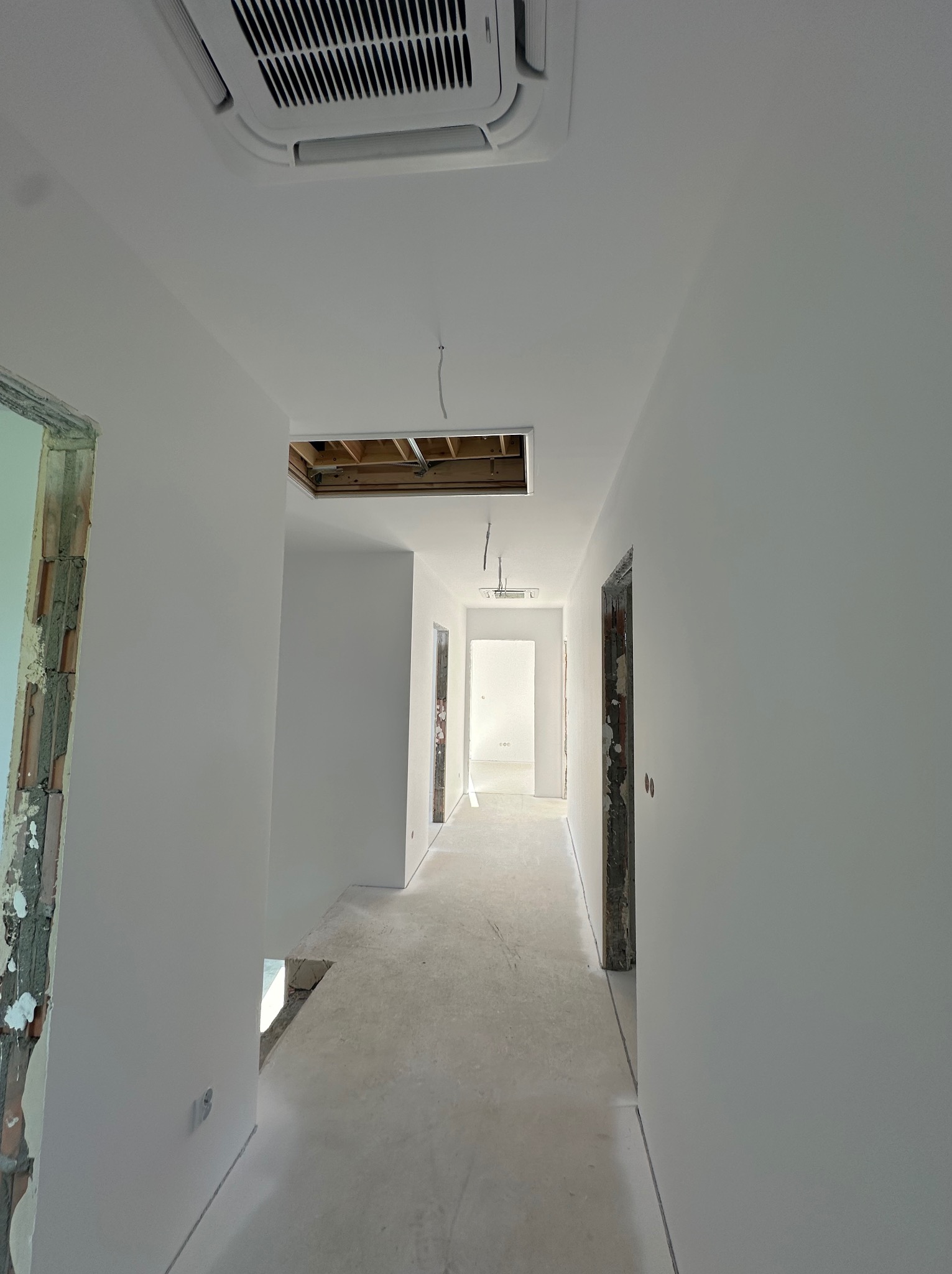 Korytarz w trakcie wykończenia: świeżo pomalowane ściany na biało, sufit z klimatyzacją i otwartym otworem, surowa futryna drzwi, betonowa podłoga.