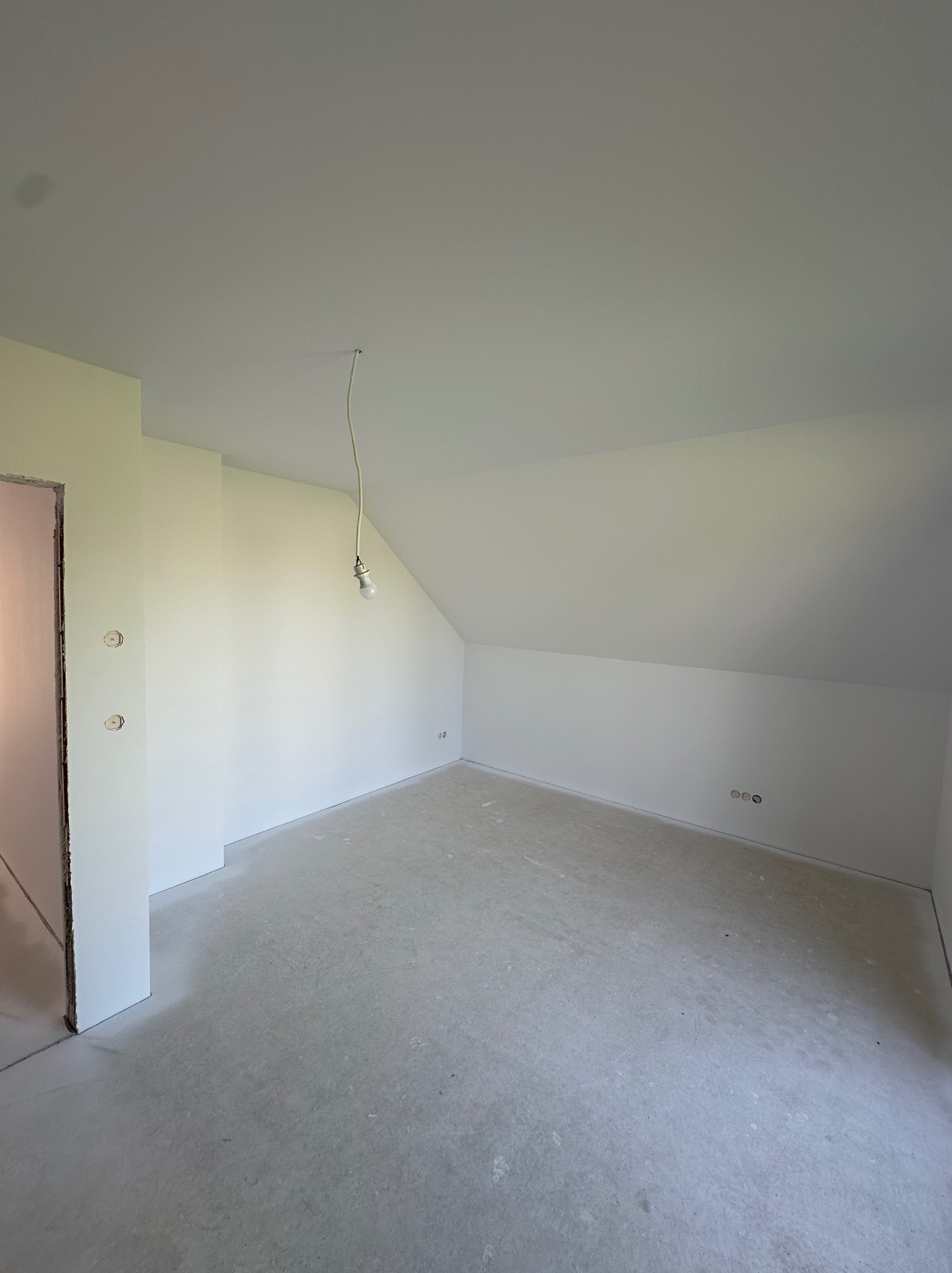 Pokój na poddaszu po gładziowaniu i malowaniu na biało. Widoczne gniazdka elektryczne i surowa podłoga. W tle ościeżnica drzwiowa.
