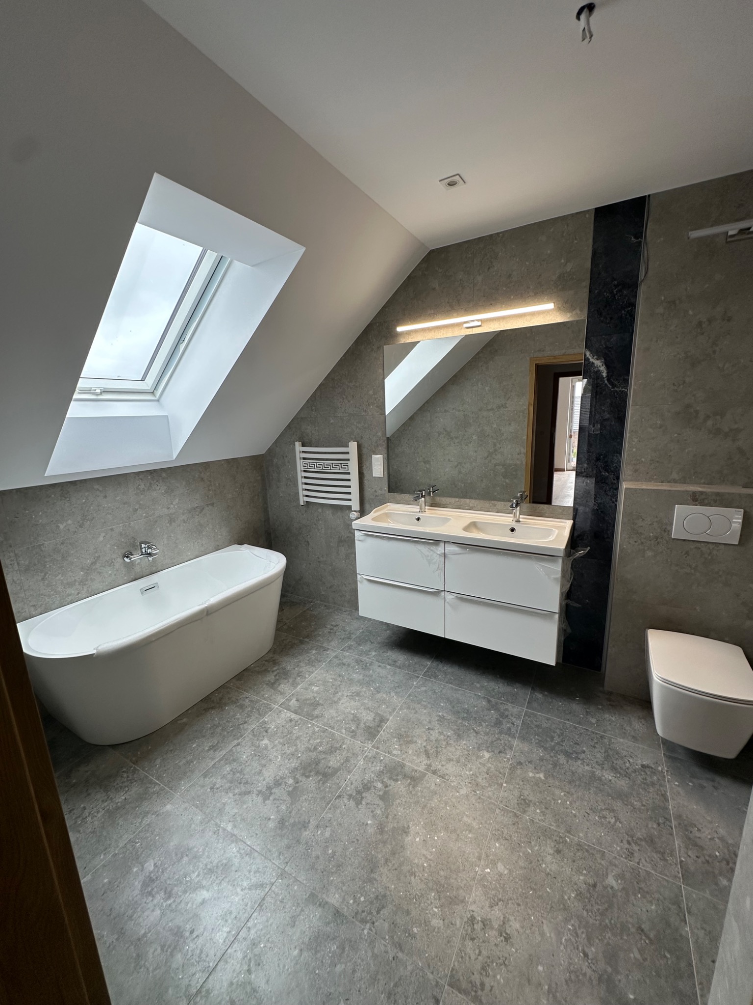 Nowoczesna łazienka na poddaszu z oknem dachowym, szarą glazurą, białą wanną wolnostojącą i szafką z umywalkami. Minimalistyczny design, oświetlenie LED.