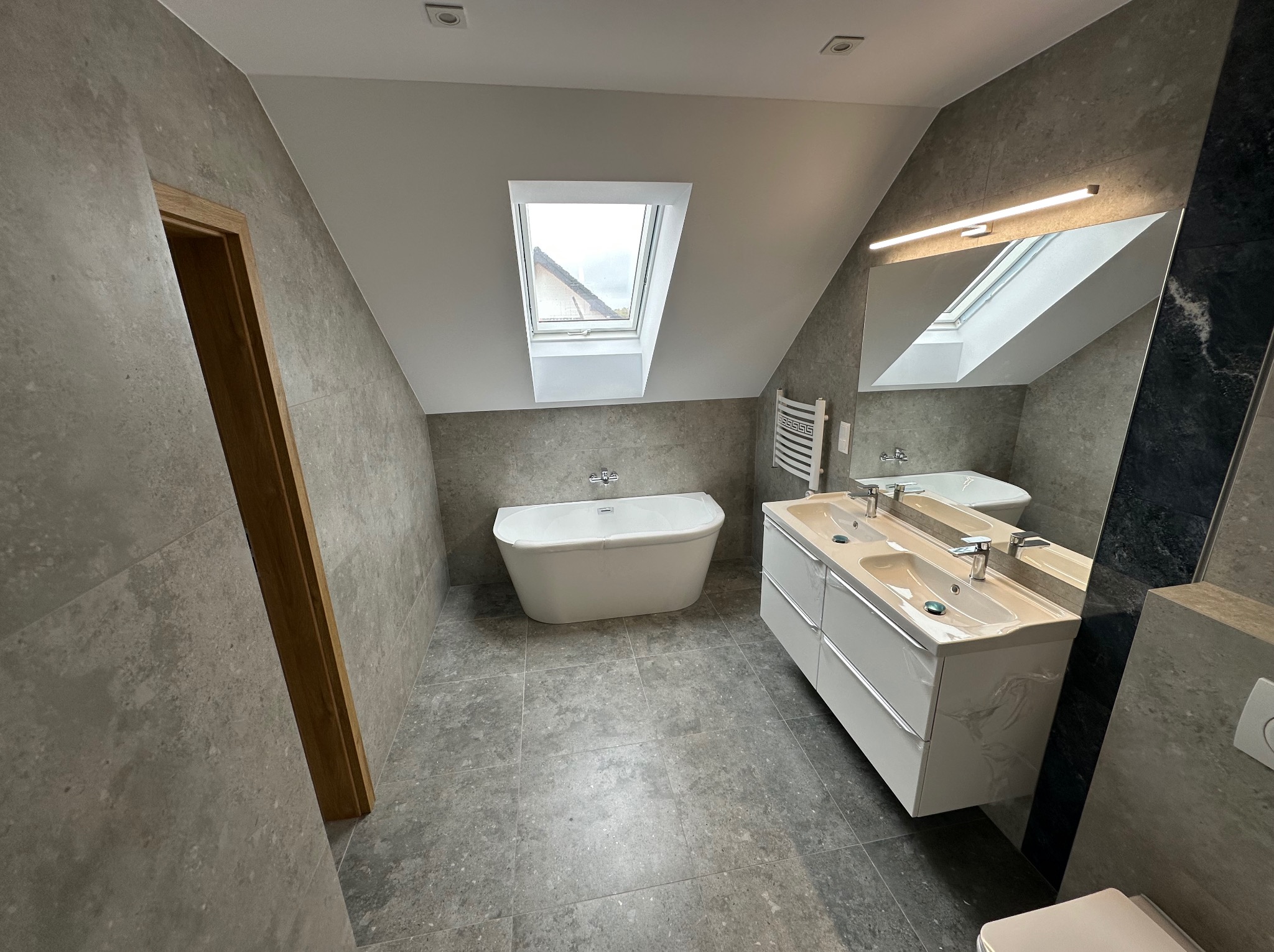 Nowoczesna łazienka na poddaszu z oknem dachowym, szarą glazurą, białą wanną i szafką z umywalkami. Minimalistyczny design, oświetlenie LED nad lustrem.
