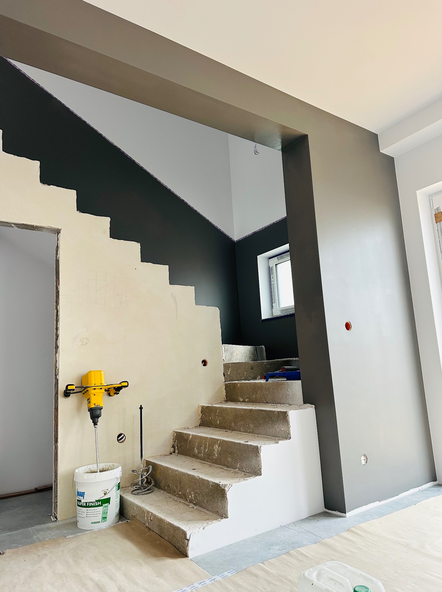 Schody w trakcie remontu: ściany częściowo pomalowane na szaro, mikser do gładzi i wiadro z gładzią. Widoczne surowe schody i przygotowane ściany.