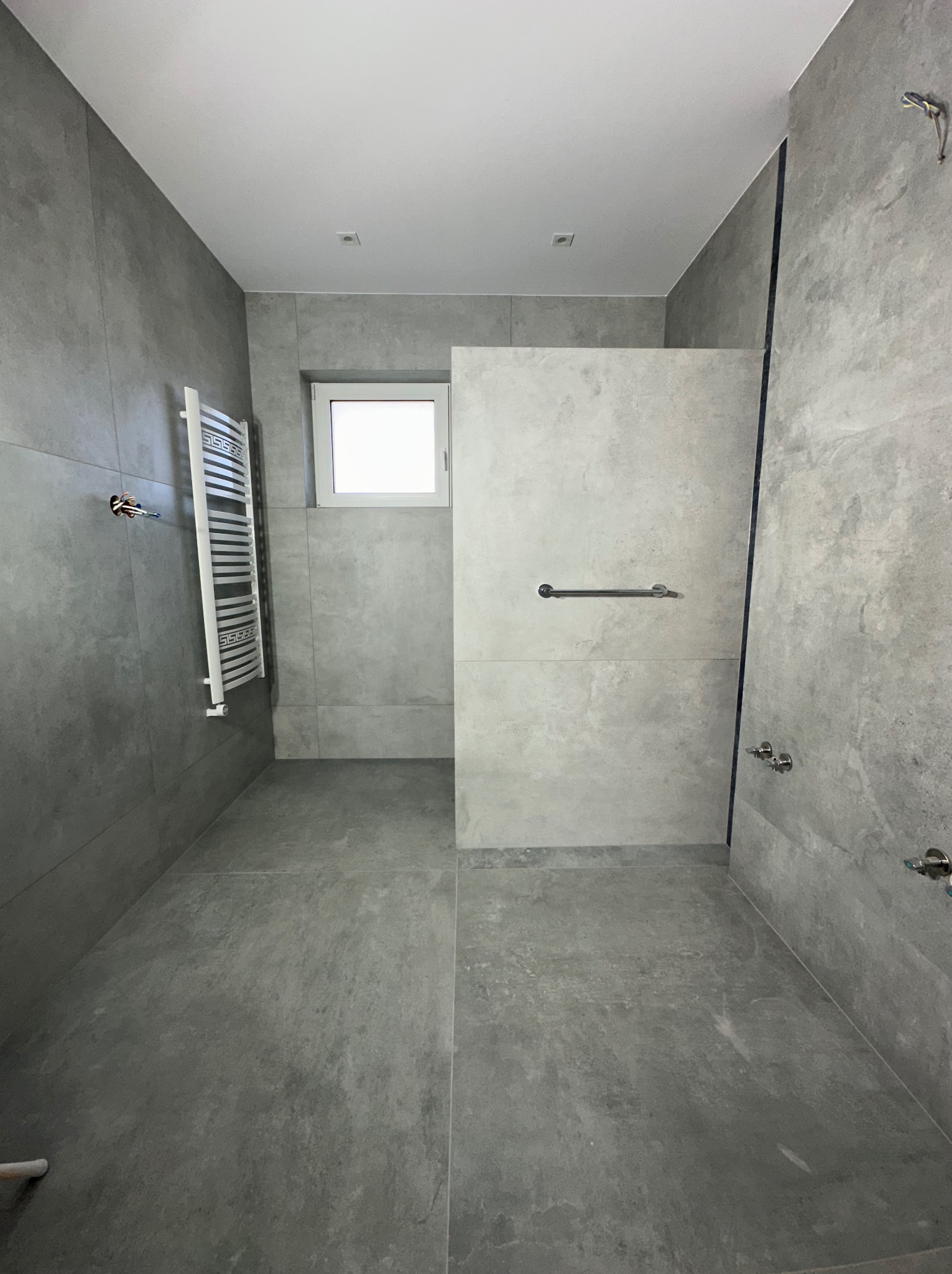 Nowoczesna łazienka w szarościach z kabiną typu walk-in, grzejnikiem drabinkowym i oknem. Minimalistyczny design, płytki imitujące kamień, chromowane elementy.