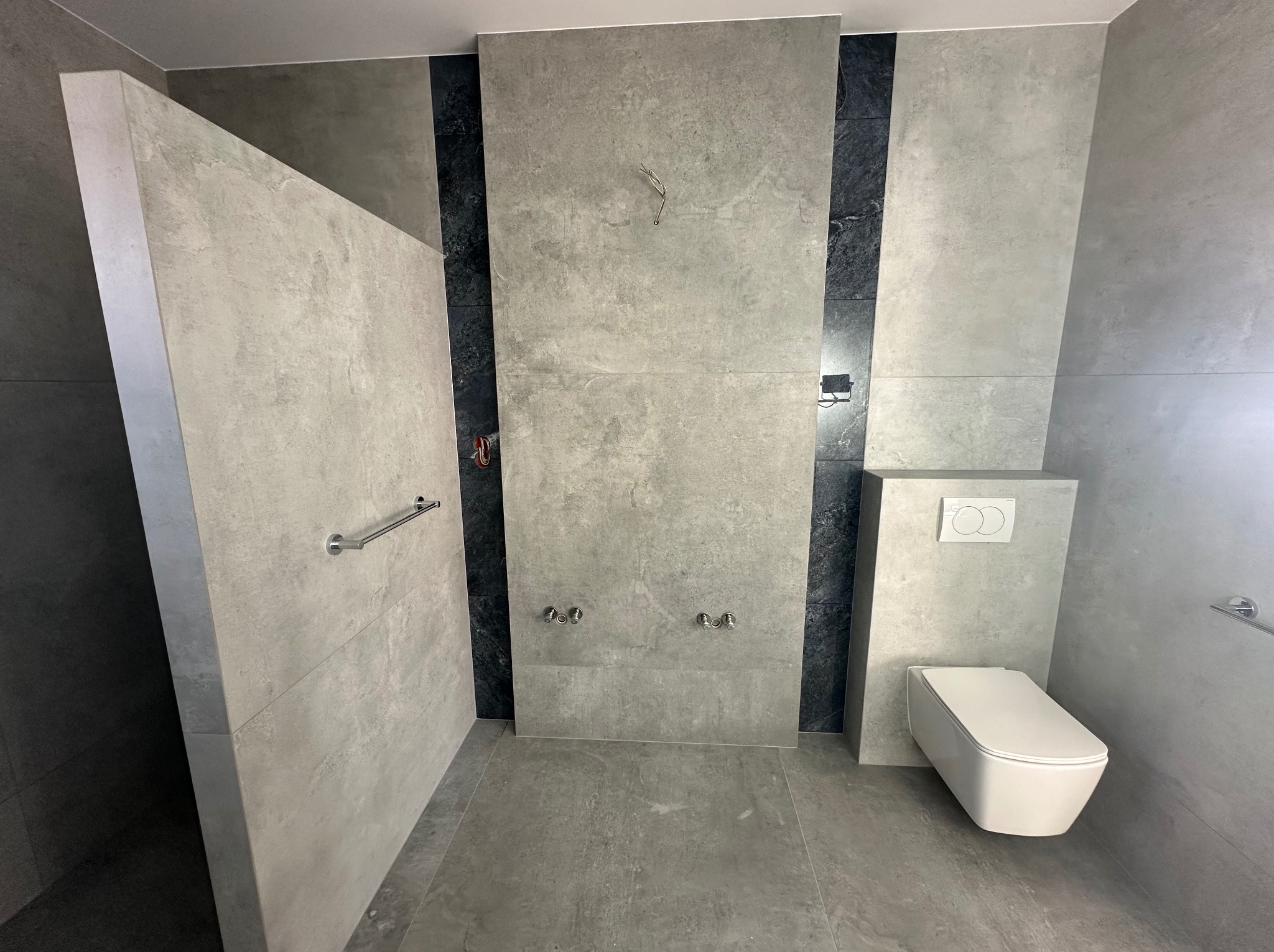 Nowoczesna łazienka w szarościach z akcentami czerni. Umywalka, toaleta i kabina prysznicowa wykończone płytkami imitującymi beton. Minimalistyczny design.