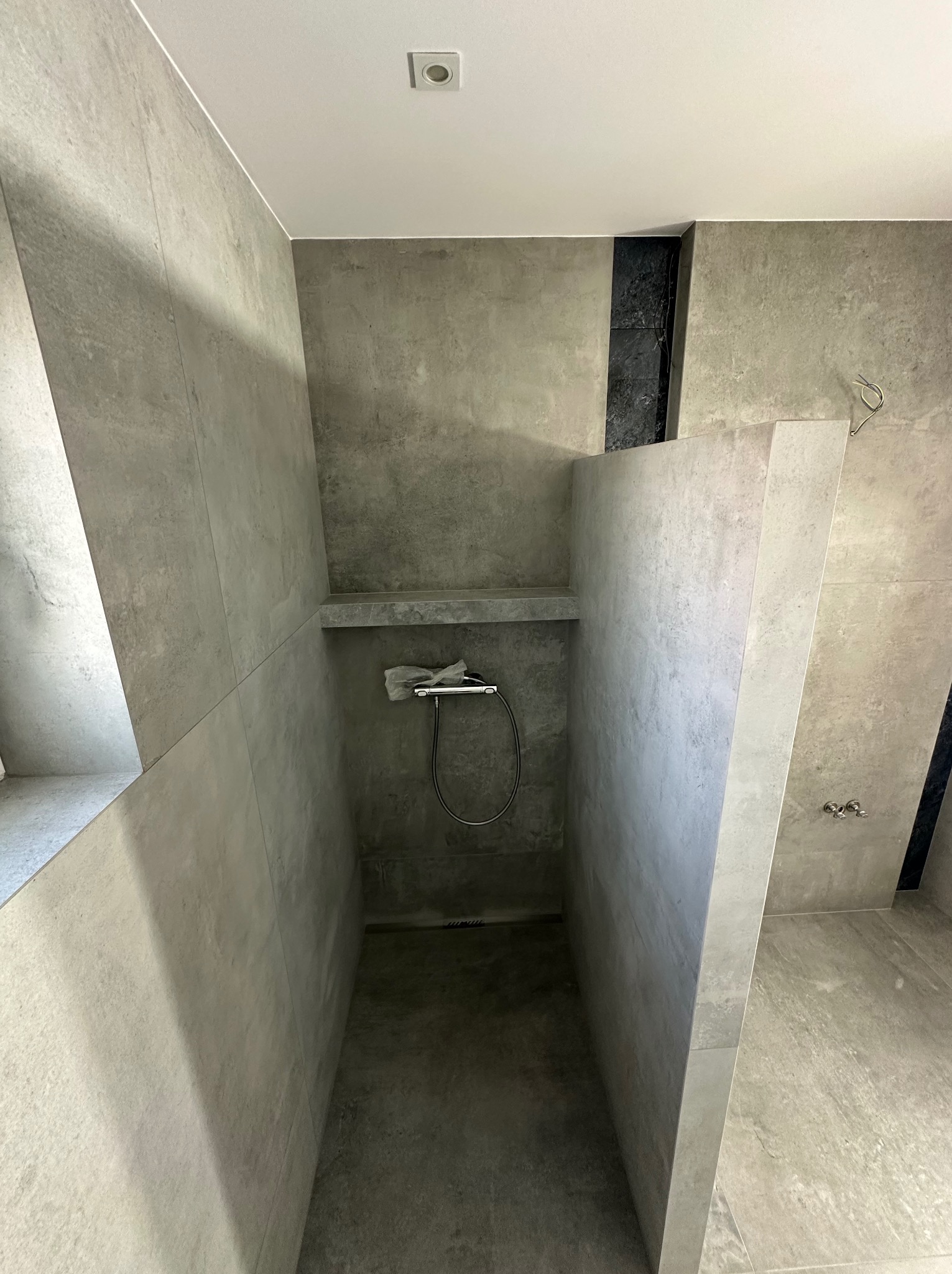 Nowoczesna kabina prysznicowa z szarymi płytkami imitującymi beton, z półką i deszczownicą w folii ochronnej. Minimalistyczny design, surowe wykończenie.