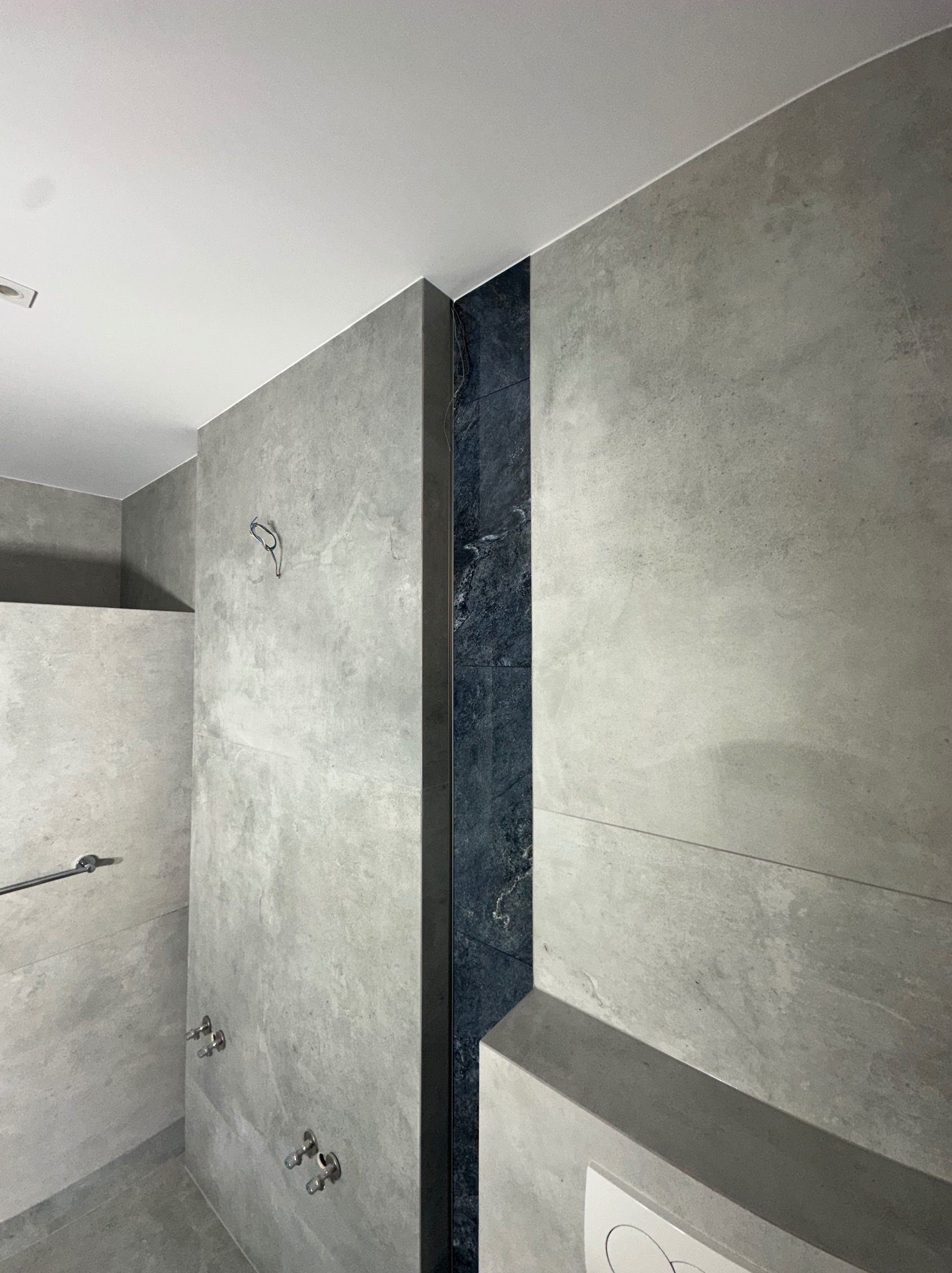 Nowoczesna łazienka z szarymi płytkami imitującymi beton i pionowym pasem z ciemnego kamienia. Widoczne baterie i uchwyt na ręcznik.