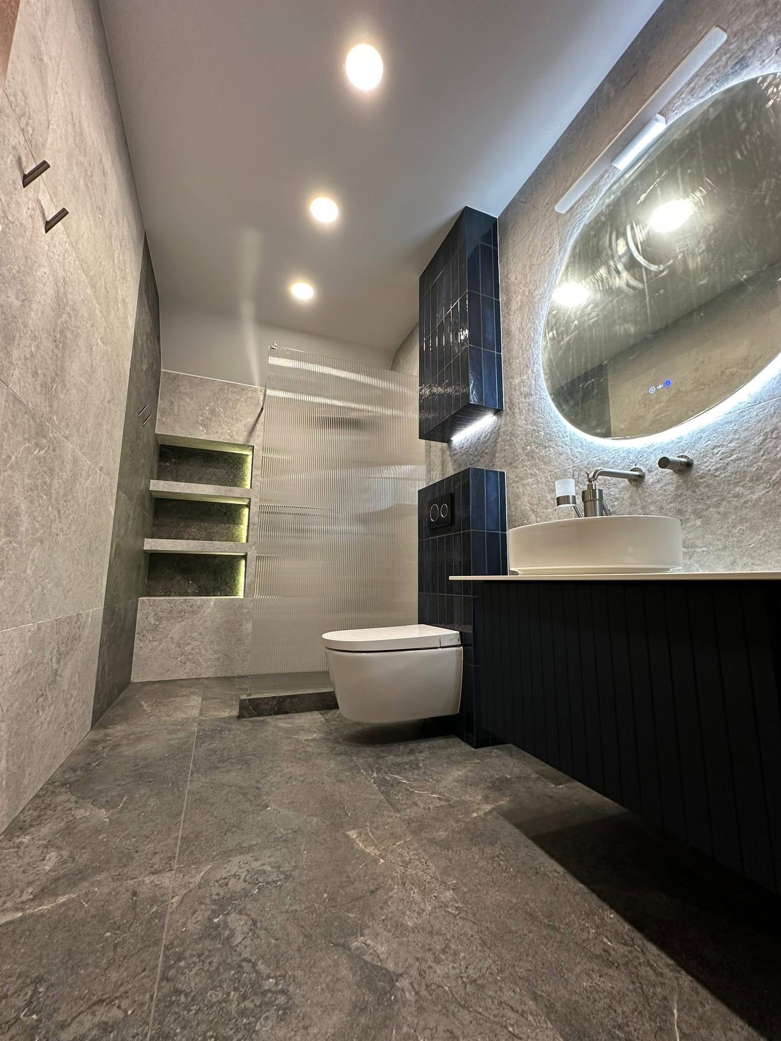 Nowoczesna łazienka z szarymi płytkami imitującymi kamień, podświetlanymi półkami i okrągłym lustrem z podświetleniem LED. Umywalka nablatowa i toaleta w zabudowie.