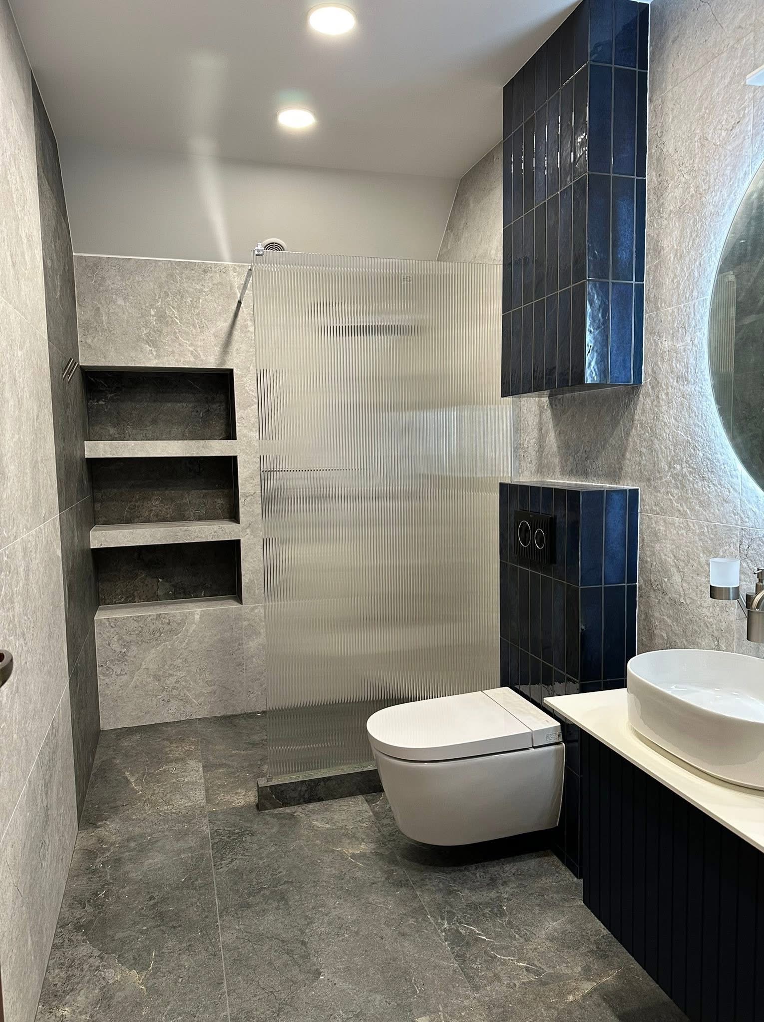 Nowoczesna łazienka z szarymi płytkami imitującymi marmur, kabiną prysznicową z ryflowanego szkła i zabudową z ciemnoniebieskich płytek typu słupek.