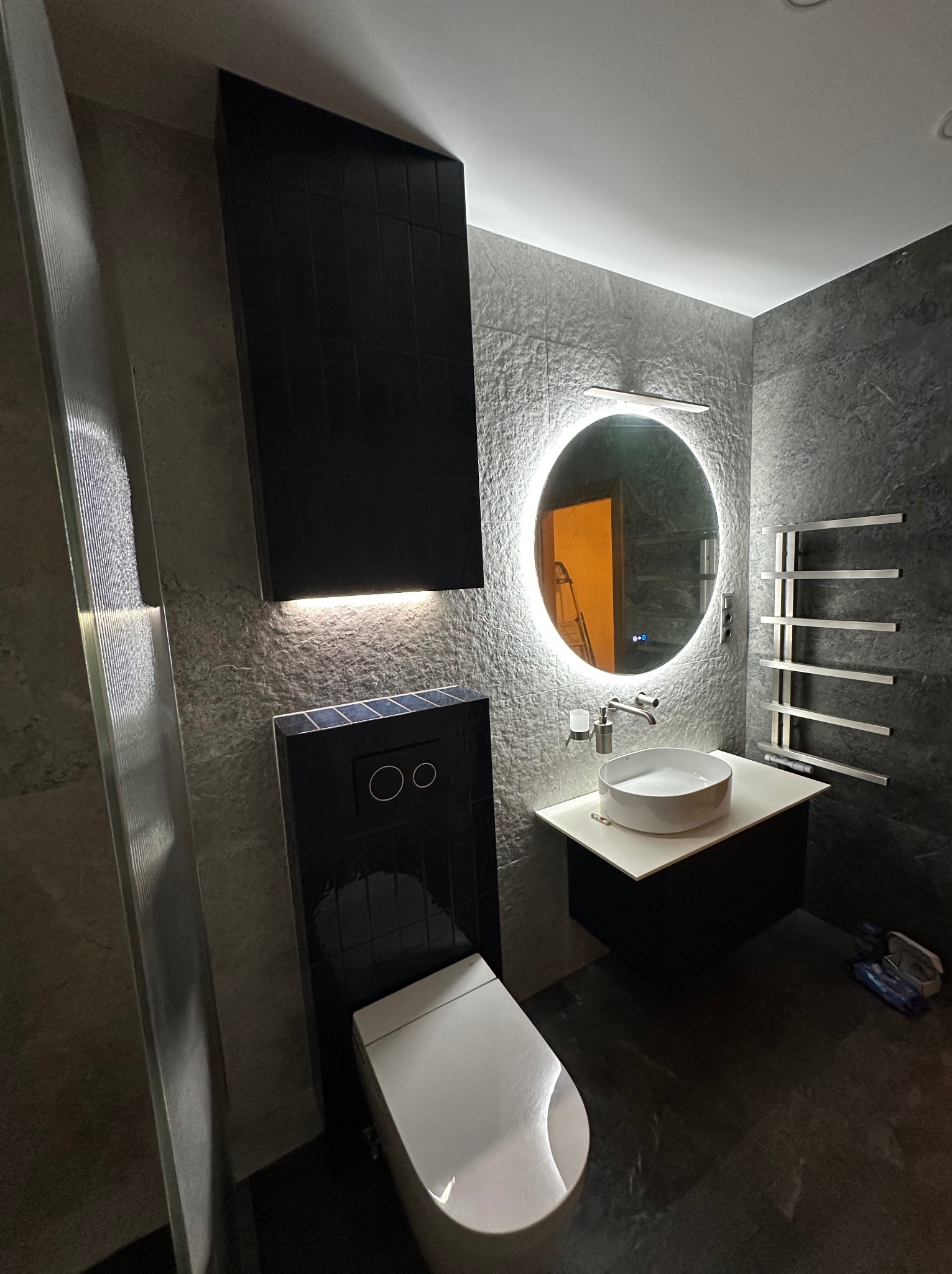 Nowoczesna łazienka w odcieniach szarości z czarnymi akcentami: podświetlane lustro, wisząca toaleta i grzejnik drabinkowy. Minimalistyczny design.