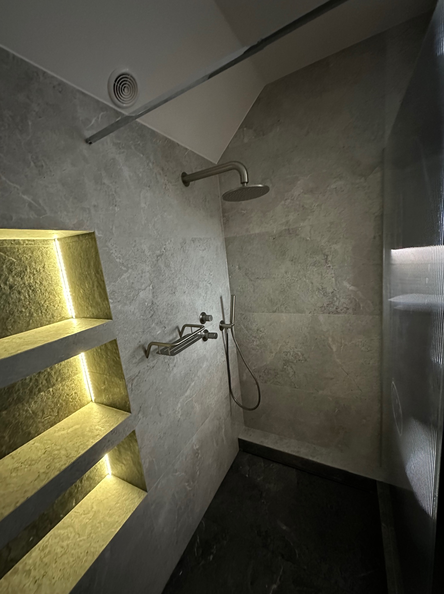 Nowoczesna kabina prysznicowa z szarymi płytkami imitującymi marmur, oświetlonymi półkami wnękowymi i deszczownicą. Minimalistyczny design, eleganckie wykończenie.
