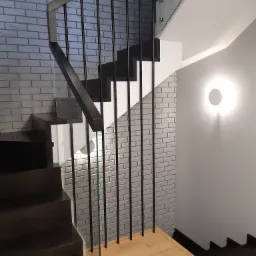 Nowoczesne schody z ciemnego drewna z czarną metalową balustradą i szklanymi panelami, zamontowane przy ścianie wyłożonej szarą cegłą, oświetlone okrągłym kinkietem.