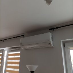 HORC SPÓŁKA Z OGRANICZONĄ ODPOWIEDZIALNOŚCIĄ - Biały klimatyzator typu split zamontowany na ścianie w jasnym pomieszczeniu, widoczna lampa podłogowa i okno z żaluzjami poziomymi.