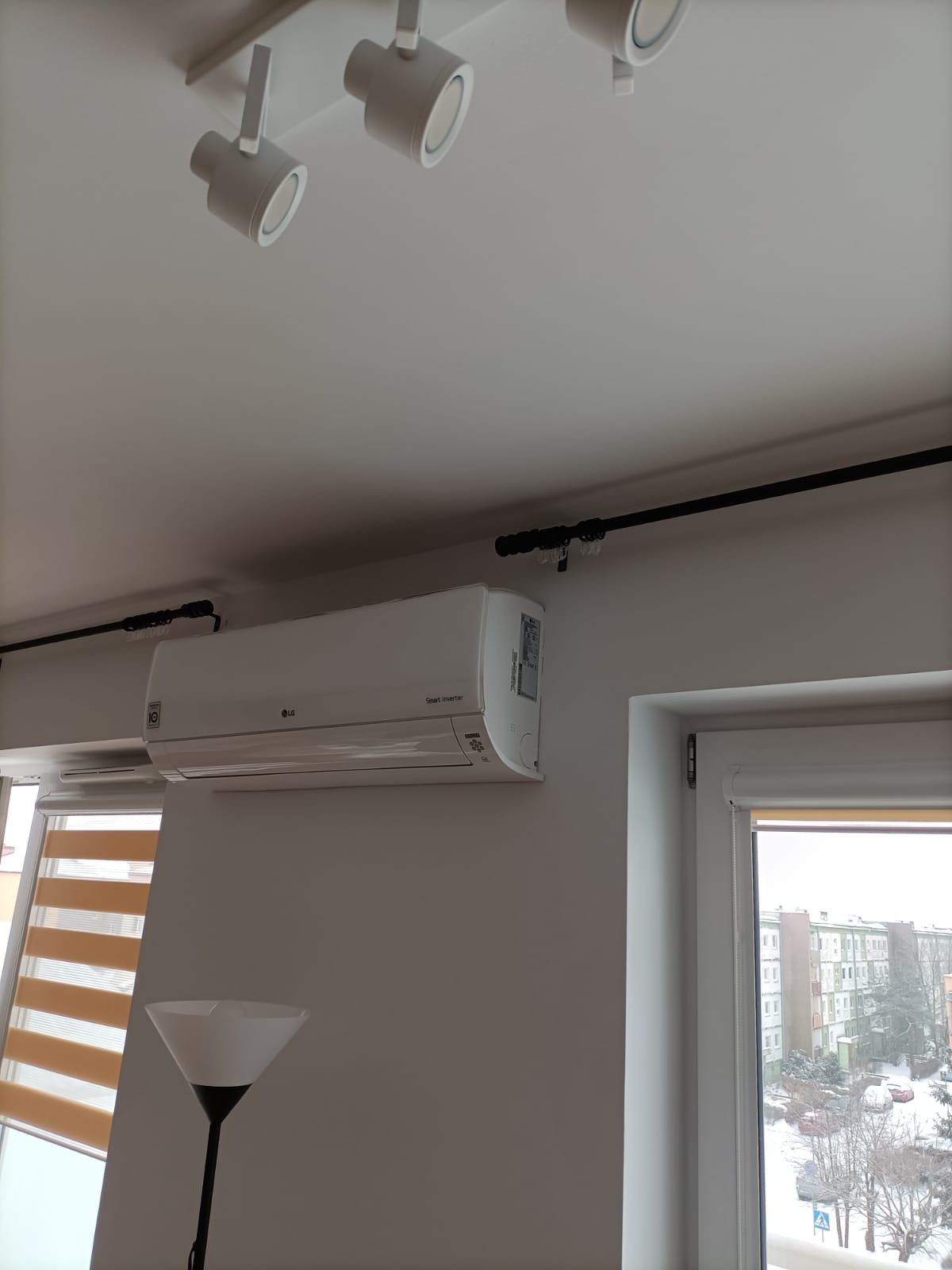 Biały klimatyzator LG Smart Inverter zamontowany na ścianie w jasnym pokoju, widoczne oświetlenie sufitowe i okno z roletami.