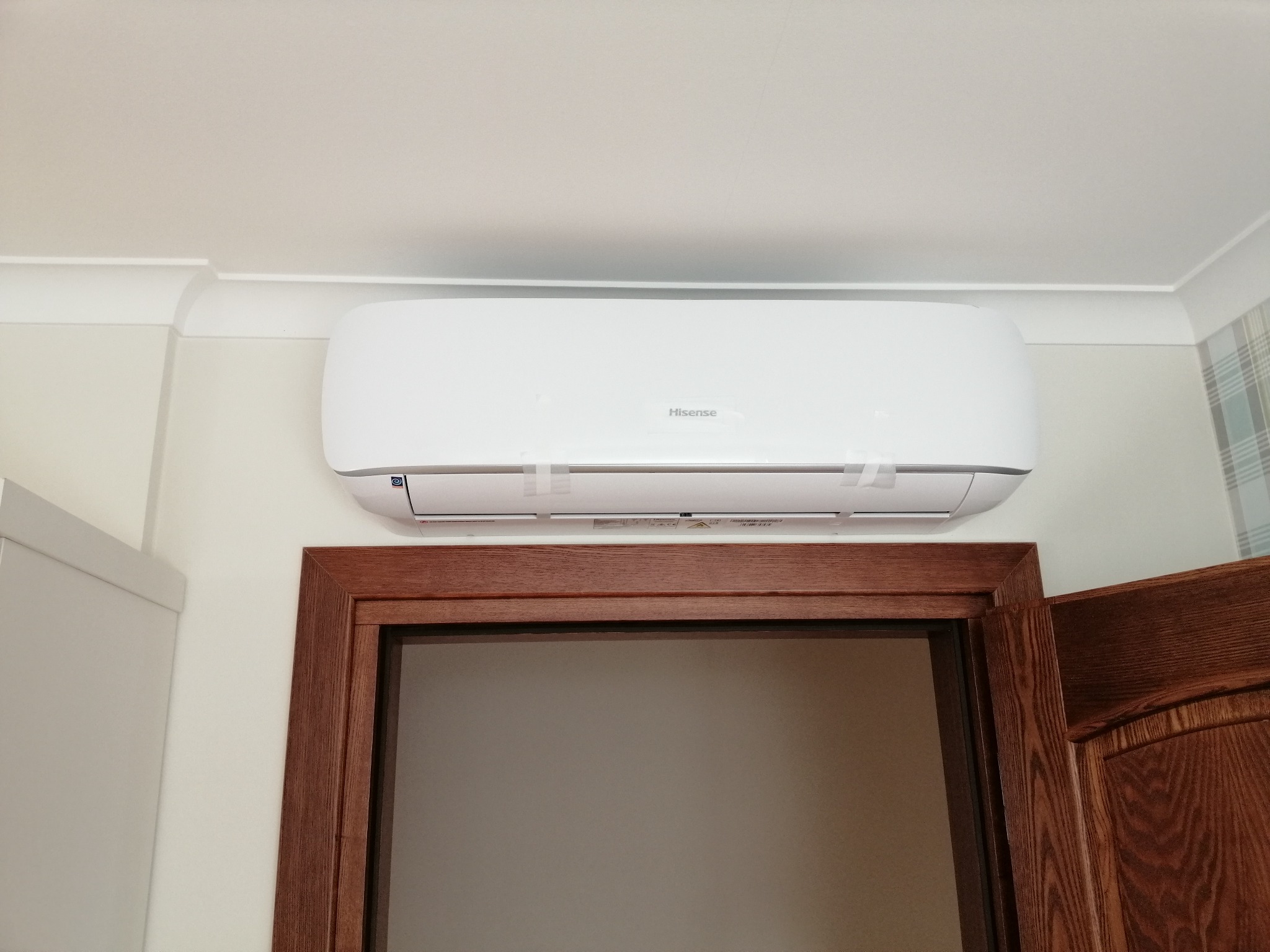 Biały klimatyzator Hisense zamontowany nad drewnianą futryną drzwiową w jasnym pomieszczeniu z dekoracyjną listwą sufitową.