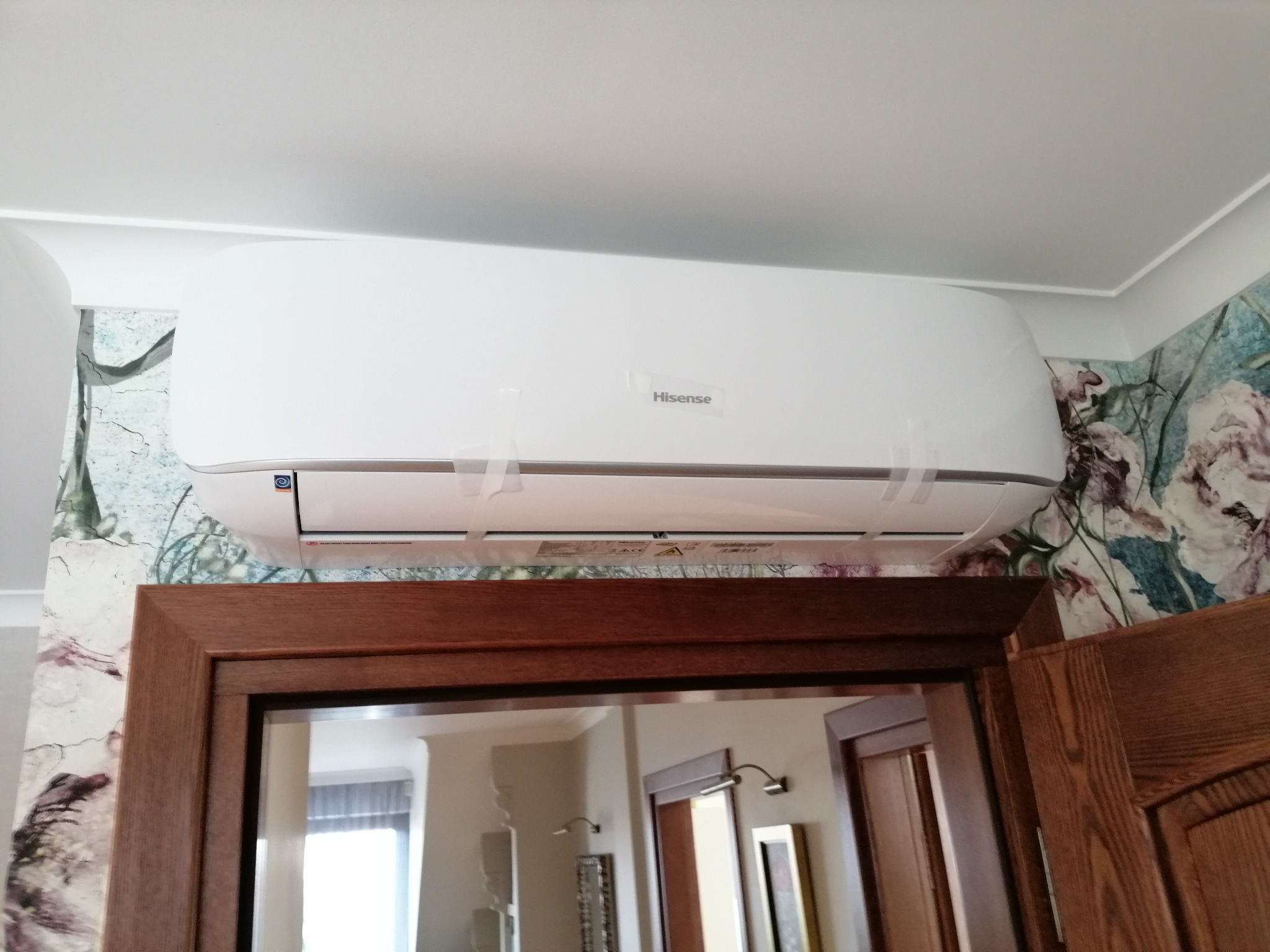 Biały klimatyzator Hisense, jeszcze z folią zabezpieczającą, zamontowany nad drewnianą futryną drzwiową w pomieszczeniu z tapetą w kwiaty i pęknięcia.