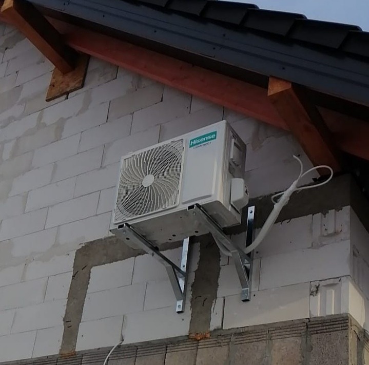 Zewnętrzna jednostka klimatyzacji Hisense zamontowana na metalowym wsporniku na ścianie budynku z bloczków betonowych, widoczne przewody i fragment dachu.