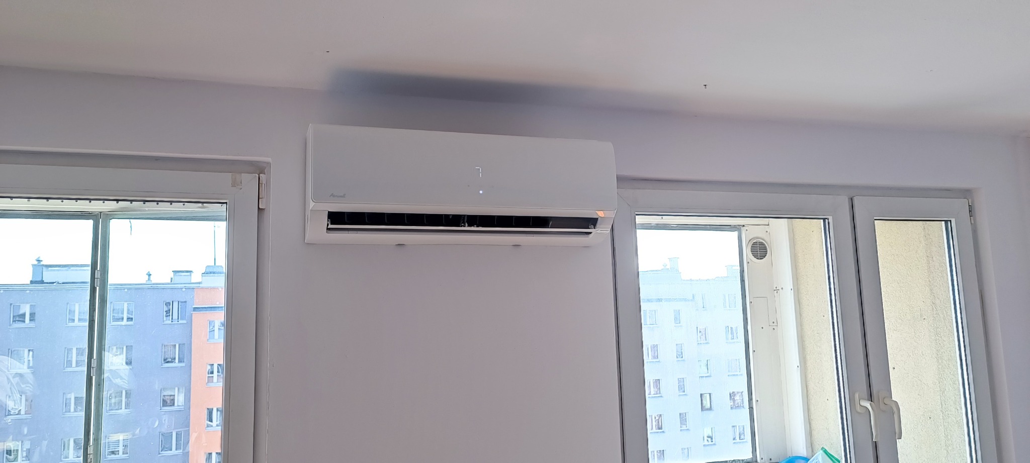 Biały klimatyzator typu split marki Airwell zamontowany na ścianie w pokoju, widoczny wyświetlacz z cyfrą 7, okna z widokiem na budynki mieszkalne.