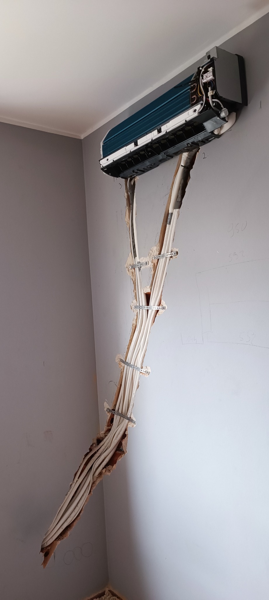 Montaż klimatyzatora ściennego z widocznymi przewodami w korycie ściennym, mocowanymi metalowymi uchwytami.