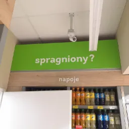 Kamera monitoringu zamontowana w sklepie w Tychach, nad regałem z napojami, pod zielonym napisem 'spragniony?'