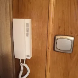 Zamontowany biały domofon z przyciskiem dzwonka na drewnianej ścianie wewnątrz mieszkania.