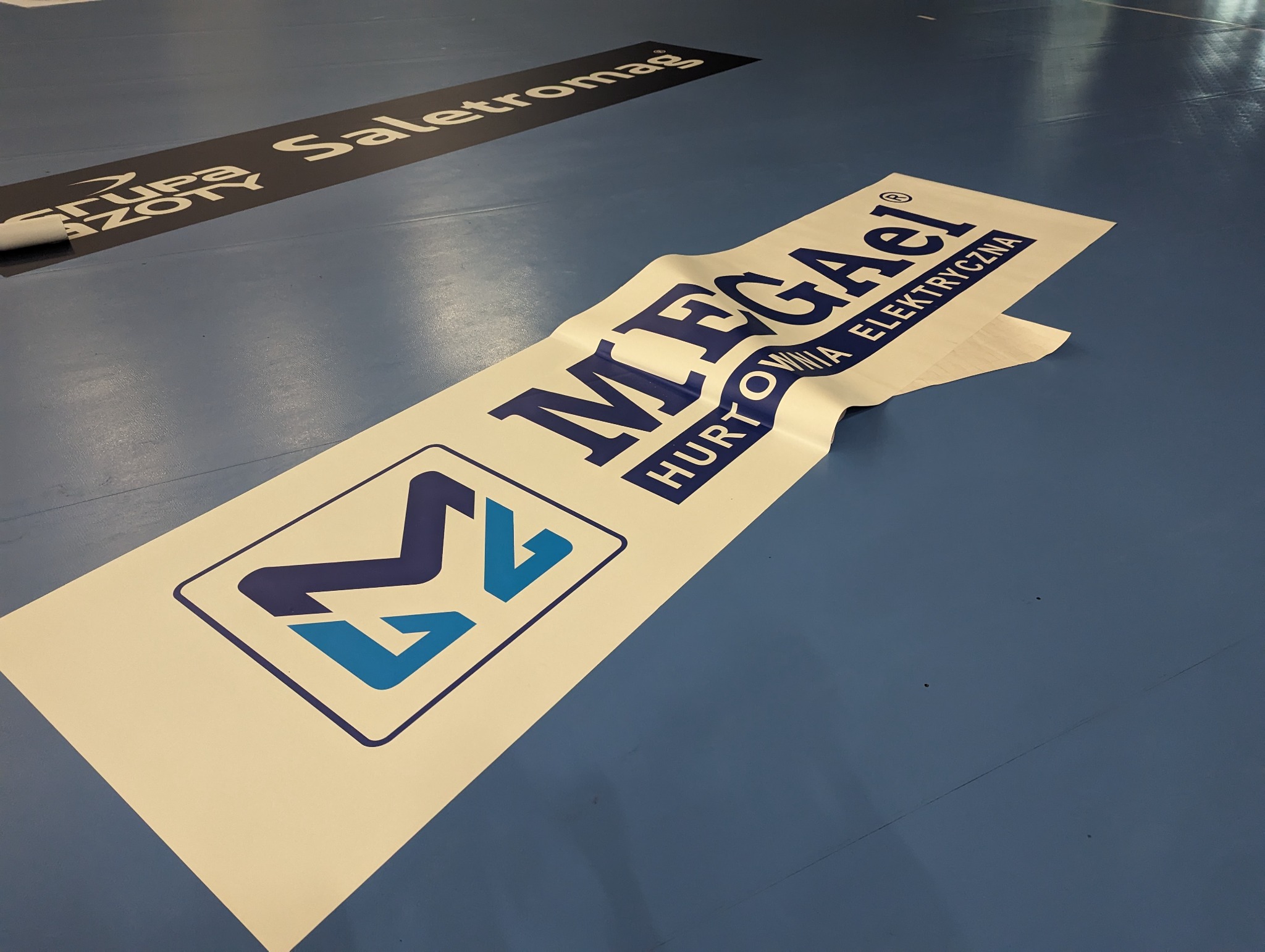 Naklejka podłogowa z logo 'MEGAel Hurtownia Elektryczna' w trakcie aplikacji, częściowo odklejona od podłoża w hali sportowej, obok widoczny fragment innej naklejki podłogowej 'Grupa Saletromag'.