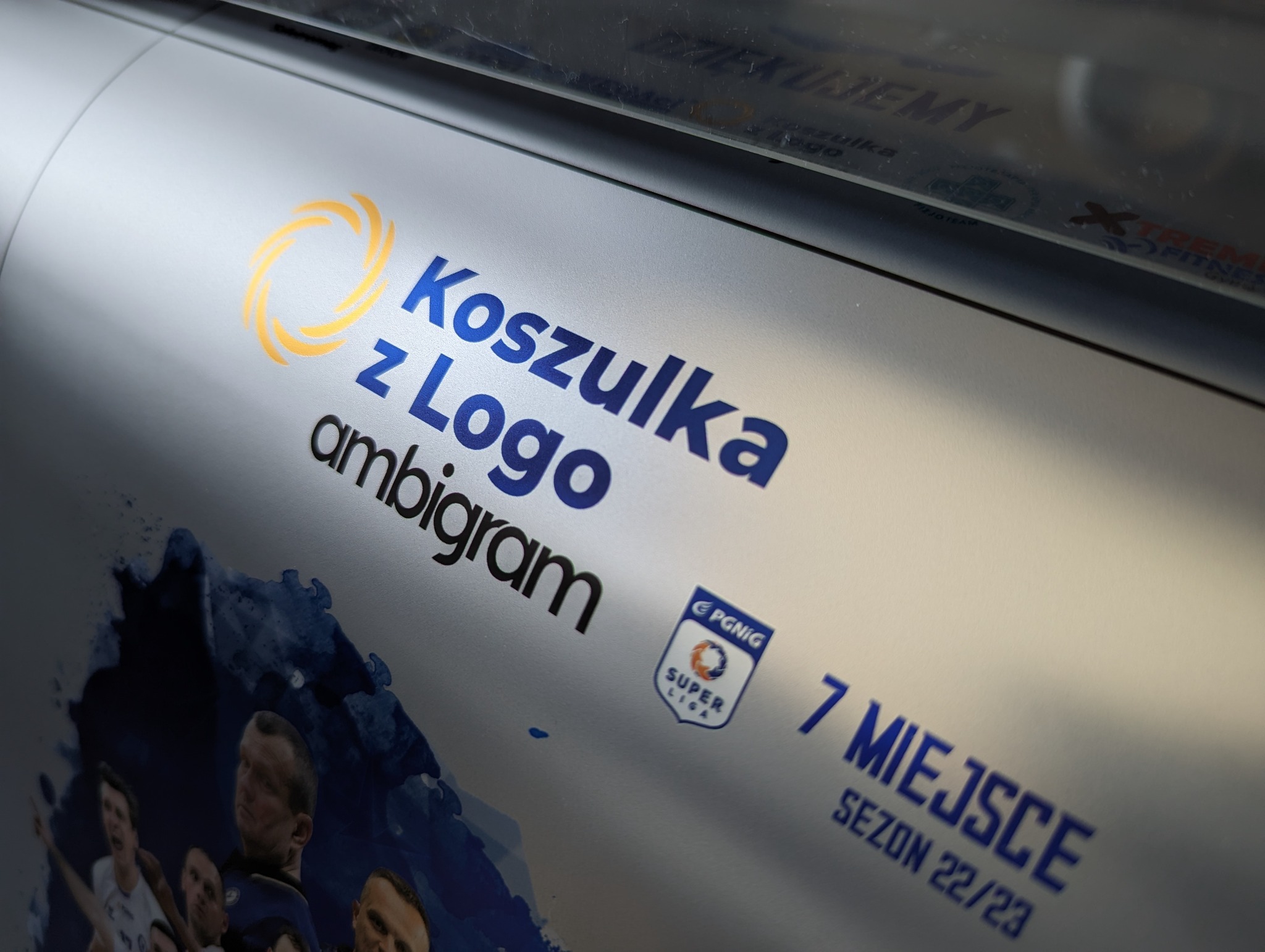Fragment wydruku z napisem 'Koszulka z Logo ambigram', logo sponsora i grafiką przedstawiającą sportowców, prawdopodobnie przygotowywany do druku na koszulce.