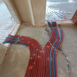 Rozdzielacz instalacji ogrzewania podłogowego z rurami PEX w kolorach czerwonym i niebieskim, przymocowanymi opaskami, widoczne ułożone pętle ogrzewania podłogowego na folii z rastrem, surowy beton...