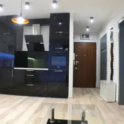 Nowoczesna kuchnia z czarnymi, lakierowanymi frontami szafek, białymi szafkami wiszącymi i drewnianą podłogą, oświetlona wiszącą lampą z pomarańczowym wnętrzem klosza.