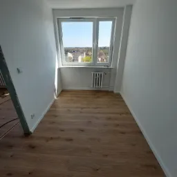 Jasne pomieszczenie w Gdańsku po remoncie, z nowymi panelami podłogowymi o wzorze drewna, świeżo pomalowanymi białymi ścianami i oknem z widokiem na miasto.